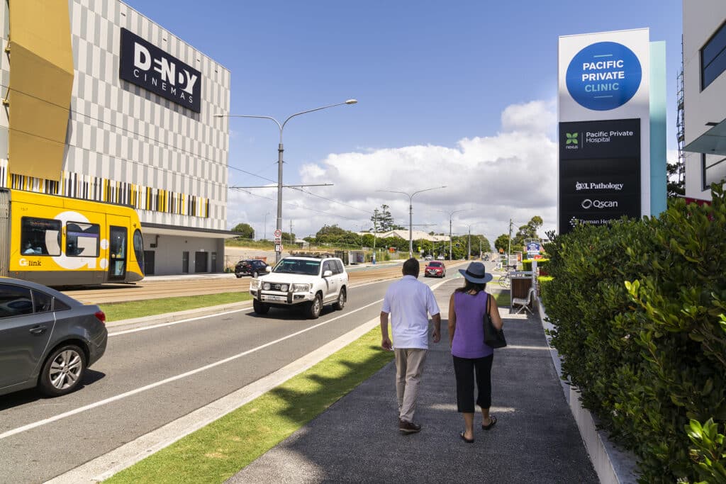 Man and lady walking toward Southport Motel & Apartments, beside road, tram, medical and Dendy cinema.