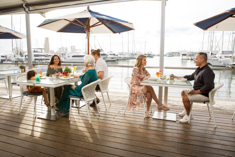People dining outside on deck under green and white umbrellas by the water